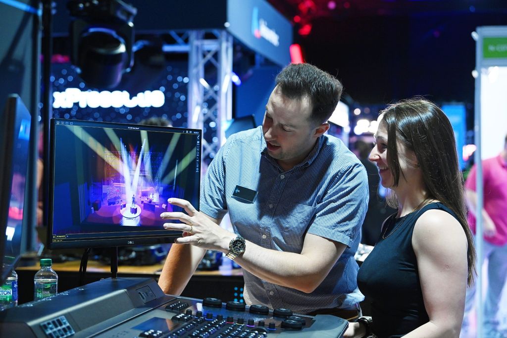 A man points to a virtual stage lighting design on a computer screen while explaining the software to a smiling woman at PLASA Focus Leeds, with a lighting control console in the foreground.