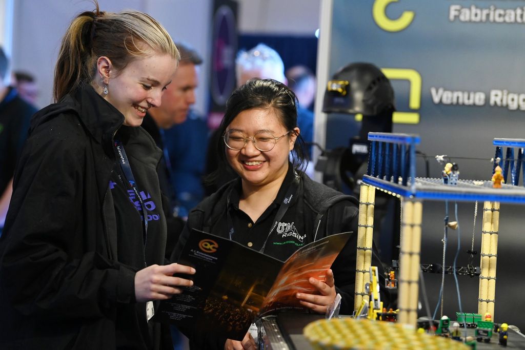Two smiling women read a brochure beside a miniature rigging and truss display at an exhibition stand during PLASA Focus Leeds