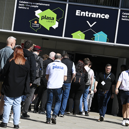 One male and four females smiling and looking at a brochure on the Area Four exhibition stand, standing beside aluminium rigging truss and flightcases at PLASA Focus Leeds.