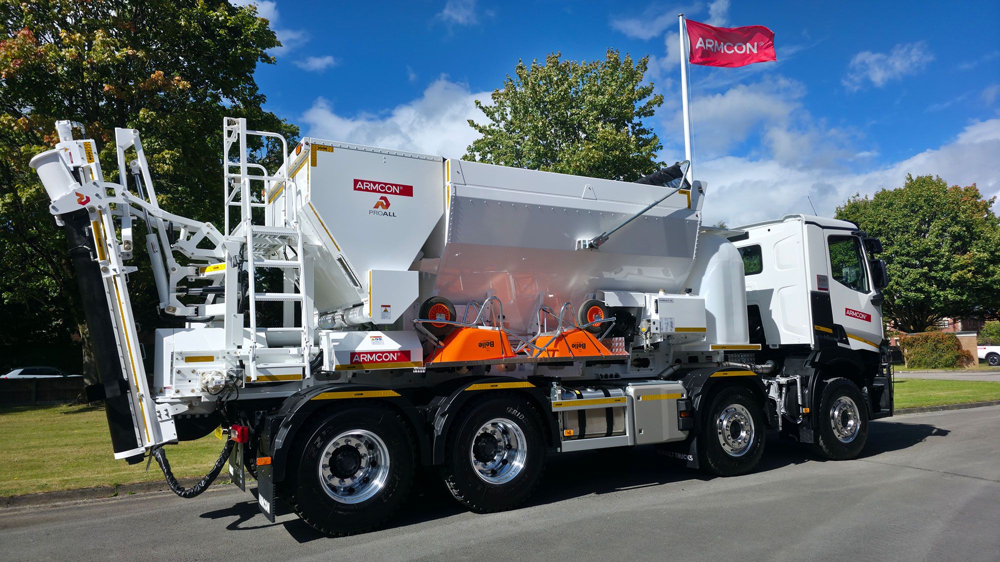Armcon ProAll L8025 volumetric concrete mixer mounted on a multi-axle truck, featuring aggregate and cement bins, rear access ladder and discharge conveyor, photographed outdoors with Armcon flag in the background