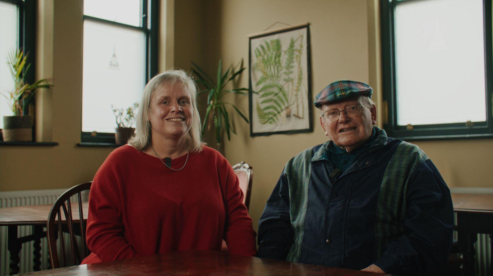 Lady wearing red jumper and older gentleman wears glasses and a flat cap and green jacket