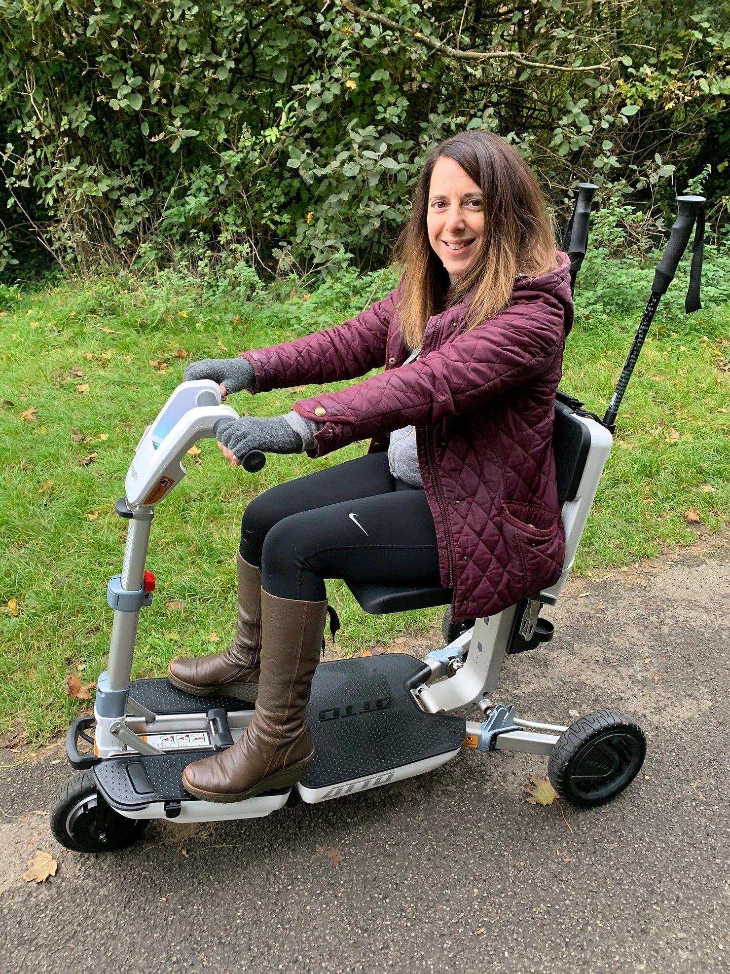 Georgina, a woman with shoulder-length brown hair, sits on a mobility scooter in front of some grass