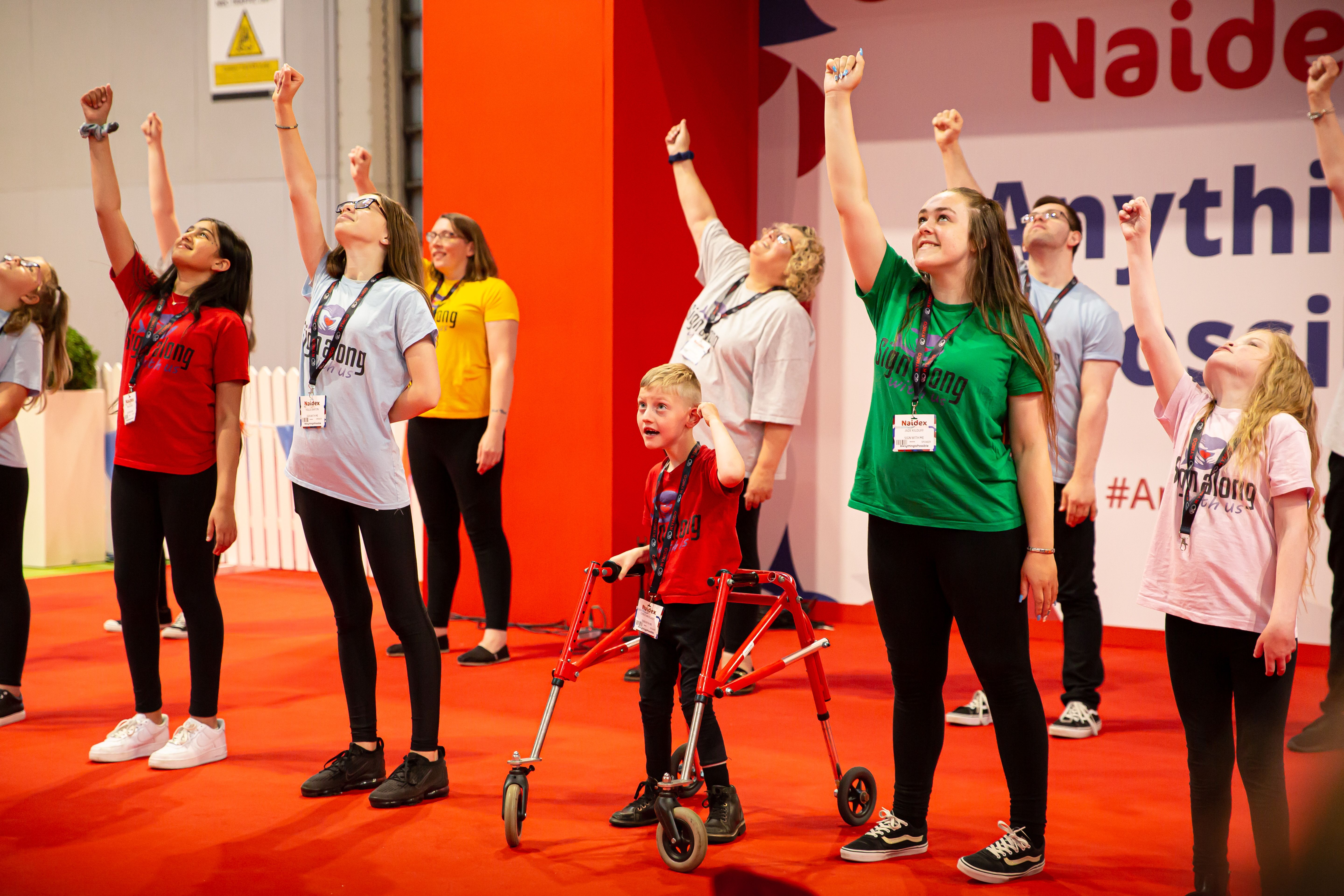 A group of children are performing a dance. They are stood up with one arm raised up high. 