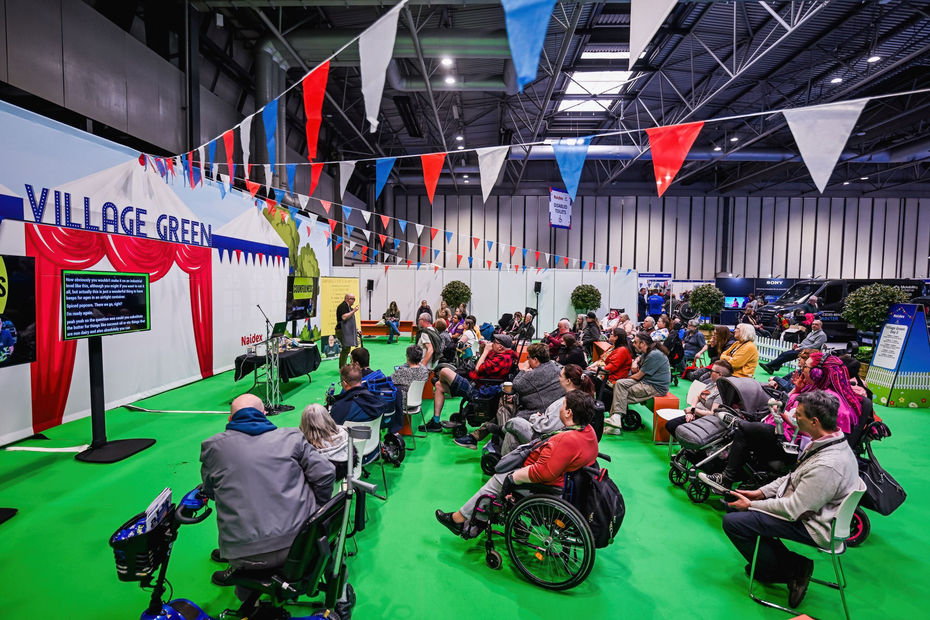 A large crowd watches a cooking demo at the village green. It is an open space with green carpet and red white and blue bunting. 