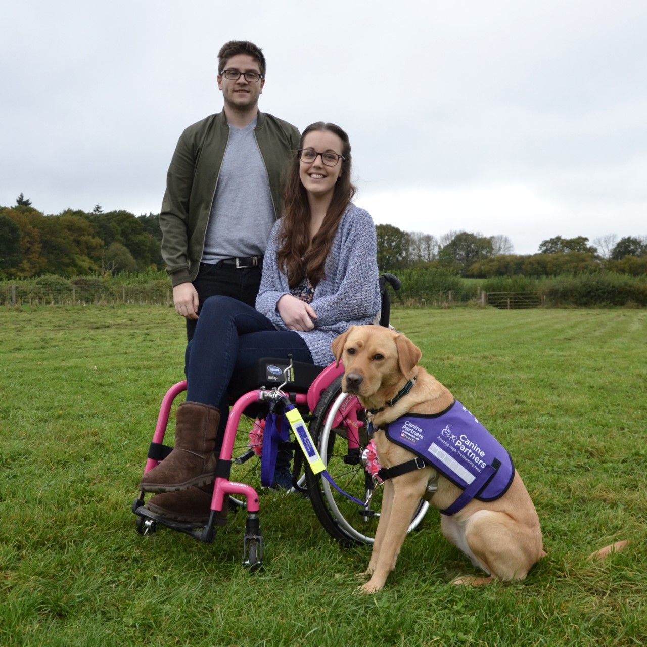 Picture of a man standing and a lady using a wheelchair, looking happy with their dog by their side.