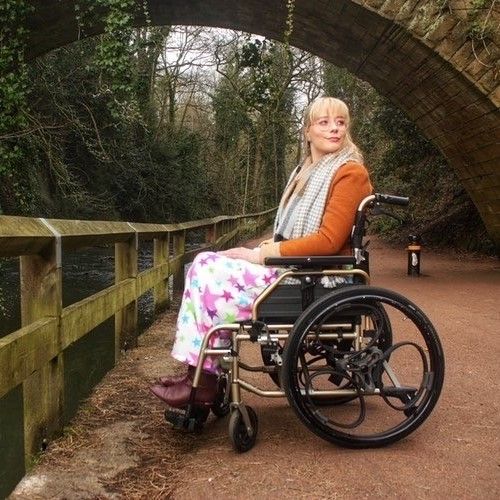 Picture of a woman using a wheelchair. She is by a river looking thoughtful.