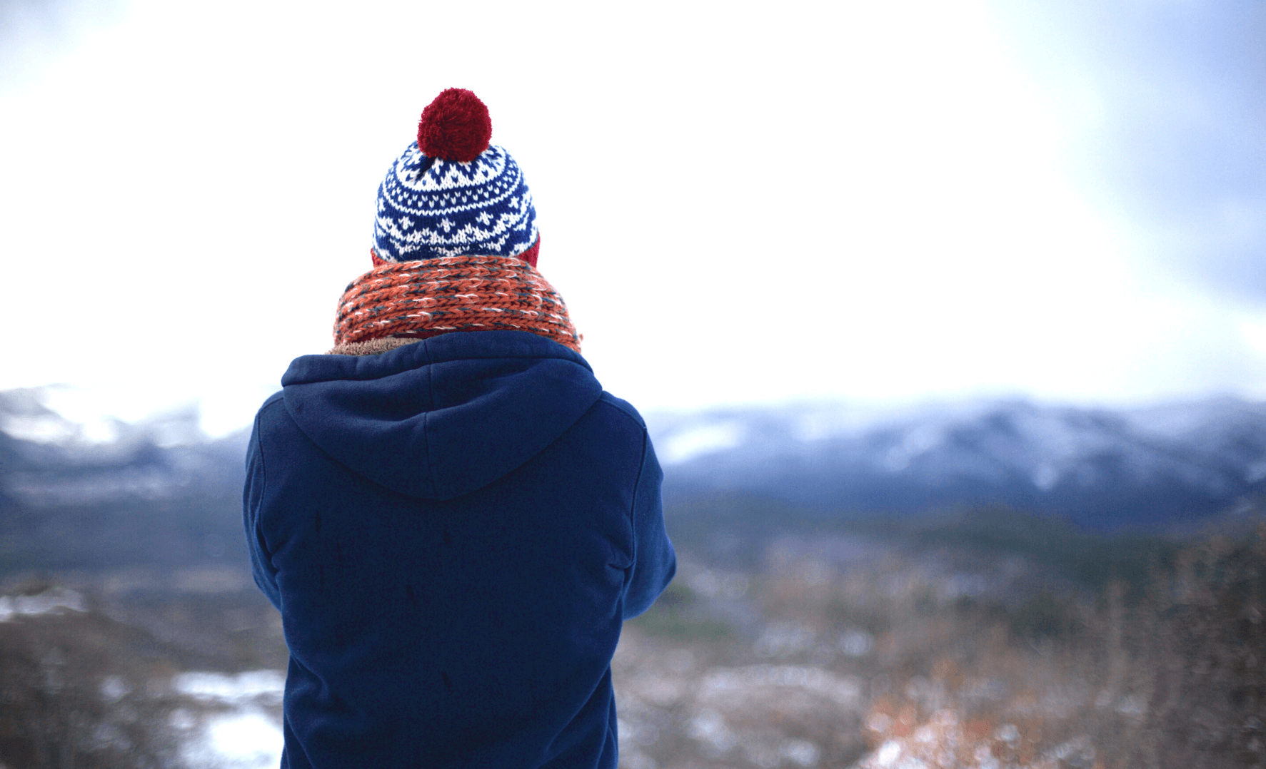 Picture of a person looking away from the camera, wearing winter clothes and a beanie  with mountains in the background.