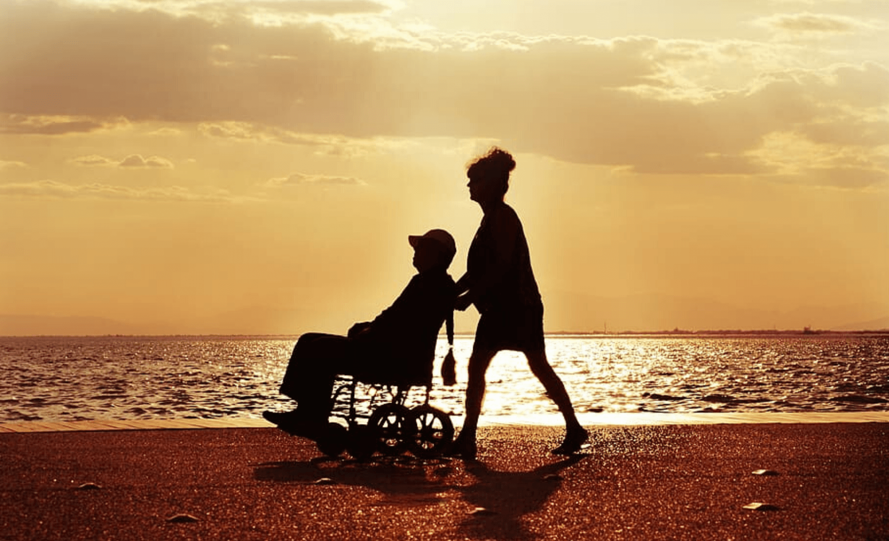 Silhouette of a lady pushing a man on a wheelchair at a beach.