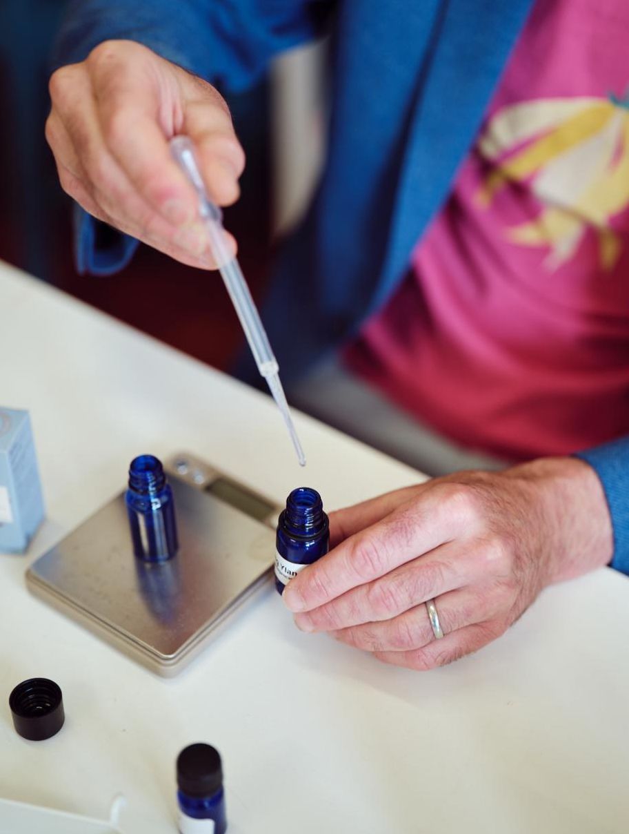 A person in a blue jacket and pink shirt uses a dropper to transfer liquid into a small blue bottle on a scale, conveying precision and focus.