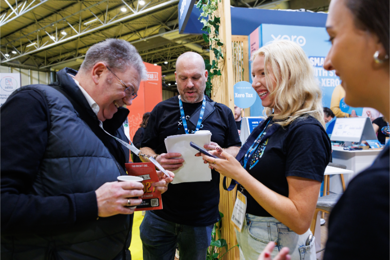 A group of people interacting at a trade show or conference booth, with one person holding a brochure and another using a smartphone.