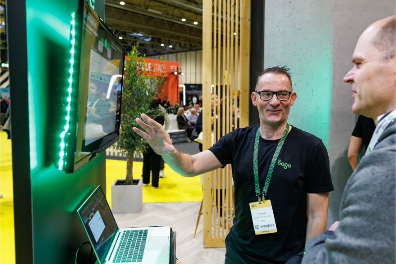 A man in a black t-shirt gestures towards a screen while speaking with another person at a trade show.