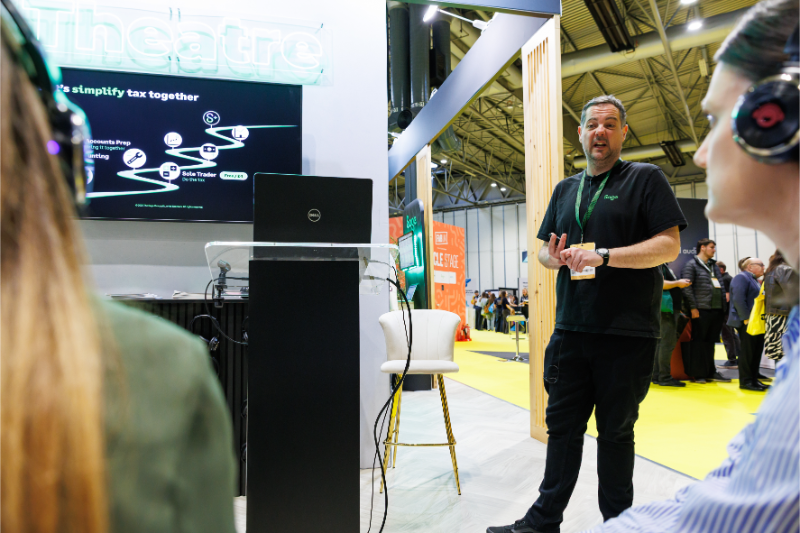 A speaker presenting at an event, with a display showing a flowchart on a screen in the background. Attendees are listening, some wearing headsets.