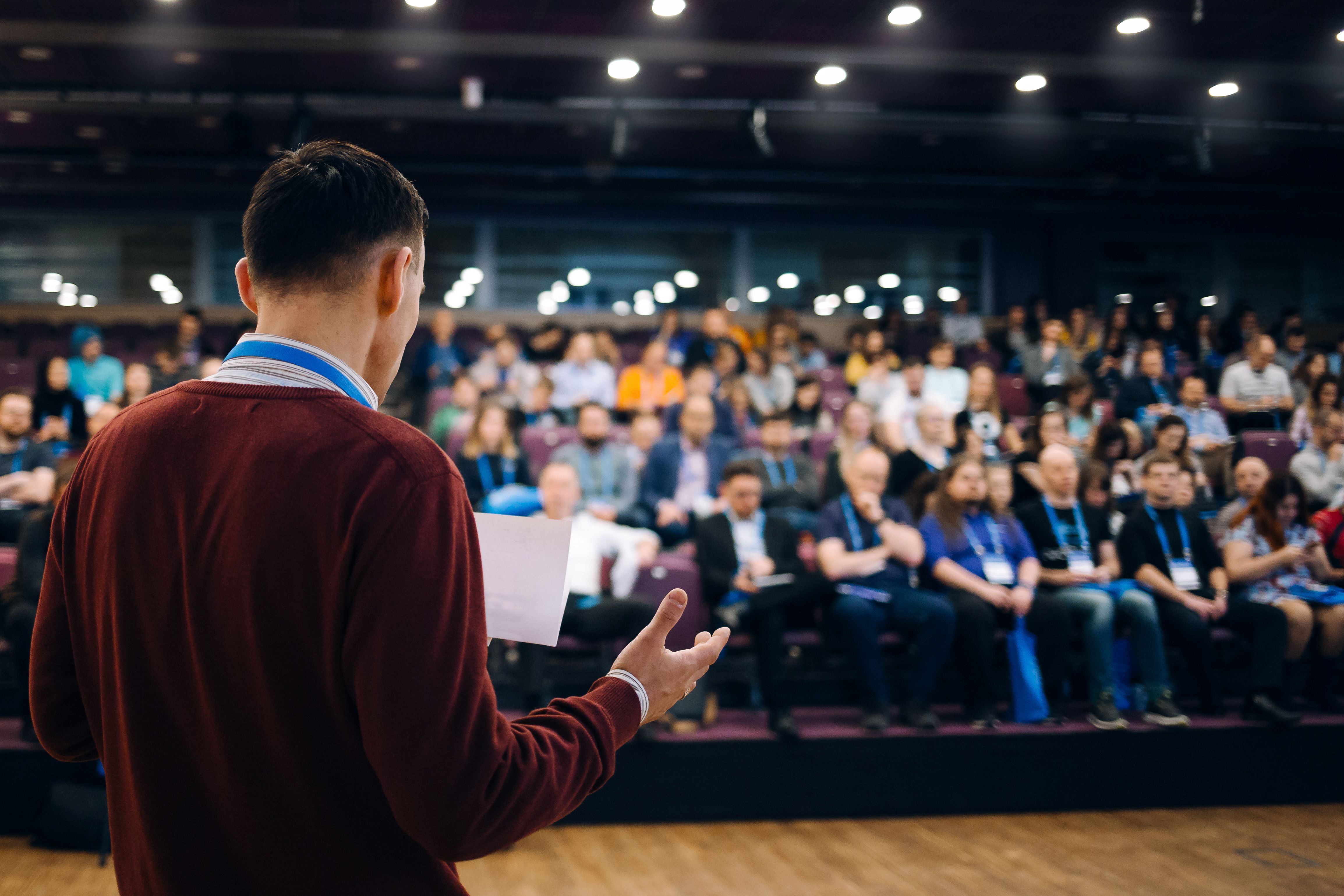 A speaker in a red sweater addresses a seated audience in a conference hall. The focus is on the speaker, with a full audience blurred in the background.