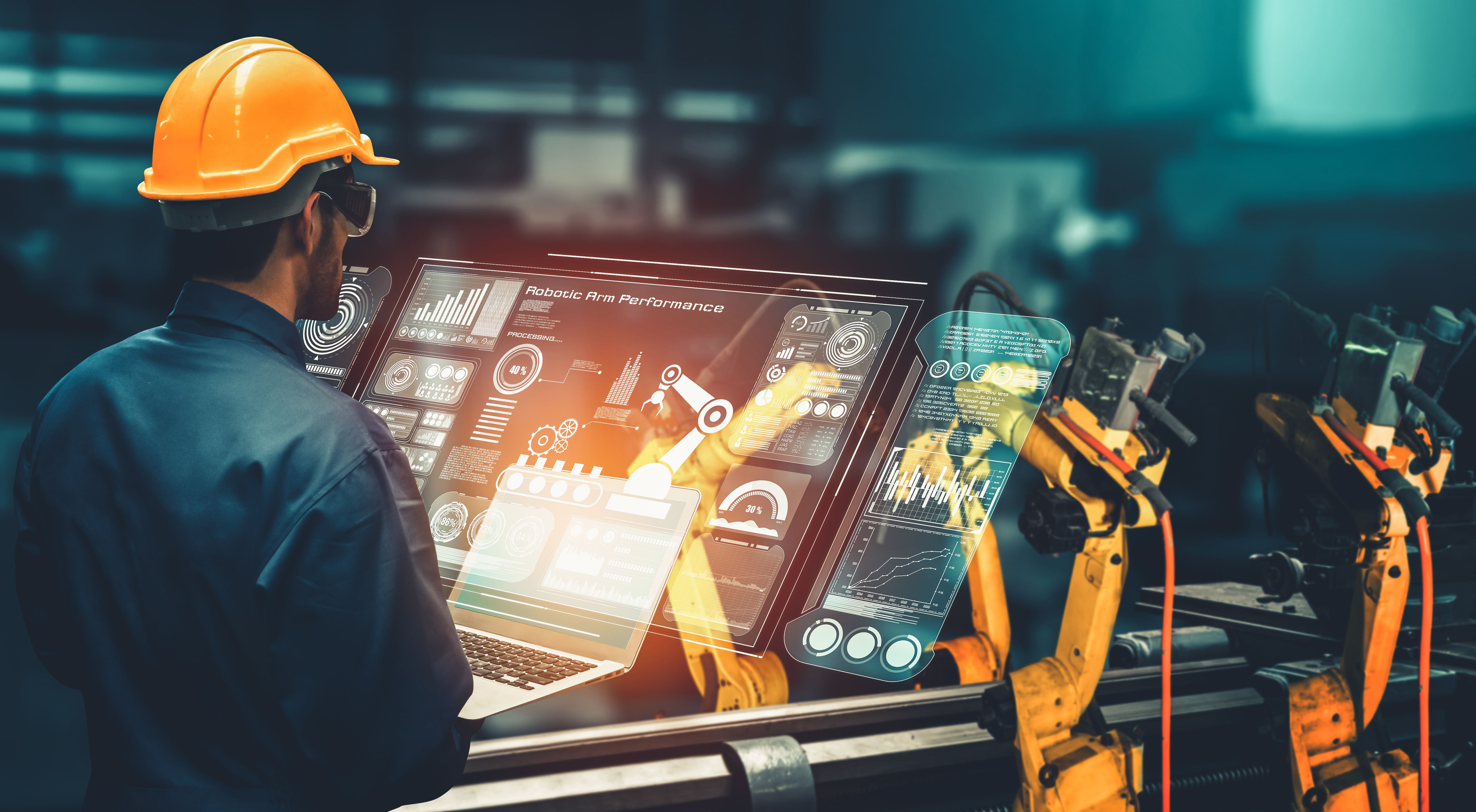 Engineer wearing a yellow hard hat operating a futuristic touchscreen interface in a factory, with robotic arms working on an assembly line. 
